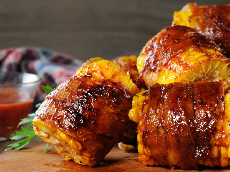 Bacon wrapped portions of corn on the cob next to a bowl of barbeque sauce